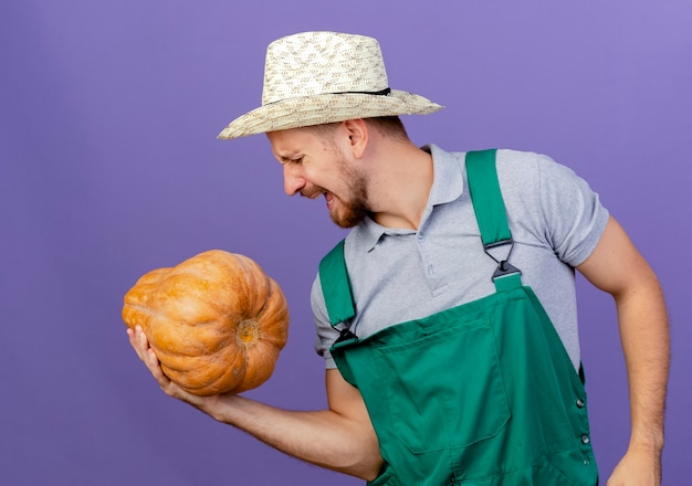 Giovane giardiniere slavo bello teso in uniforme e cappello che tiene la zucca butternut con una mano con gli occhi chiusi