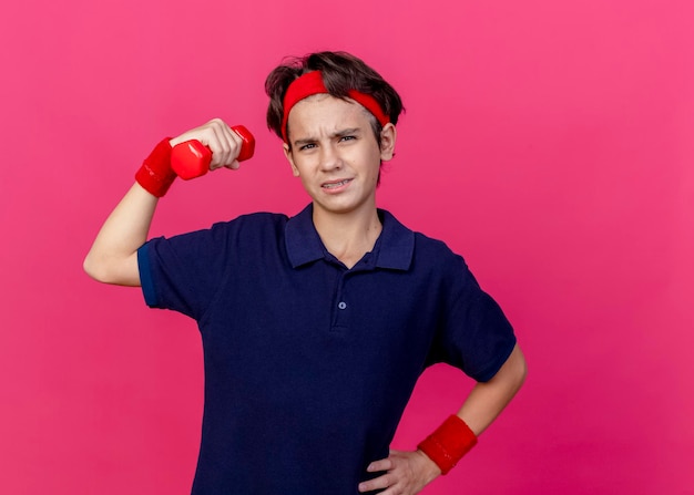 Strained young handsome sporty boy wearing headband and wristbands with dental braces keeping hand on waist raising up dumbbell looking at front isolated on pink wall with copy space