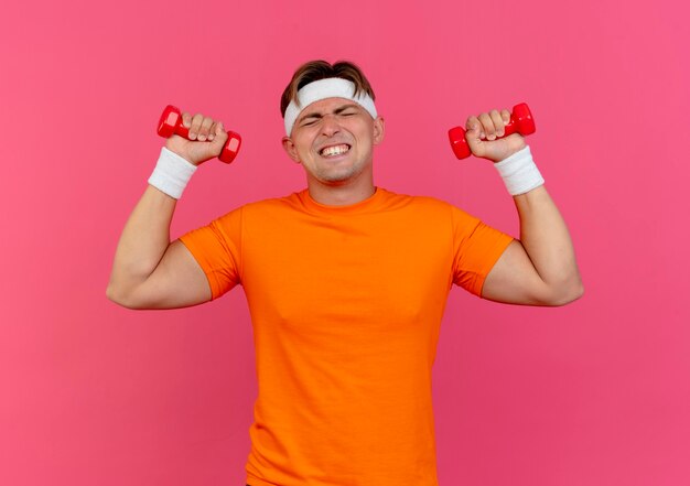 Strained young handsome sporty man wearing headband and wristbands raising dumbbells with closed eyes isolated on pink wall