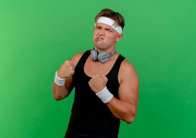 Strained young handsome sporty man wearing headband and wristbands with headphones on neck clenching fists isolated on green wall
