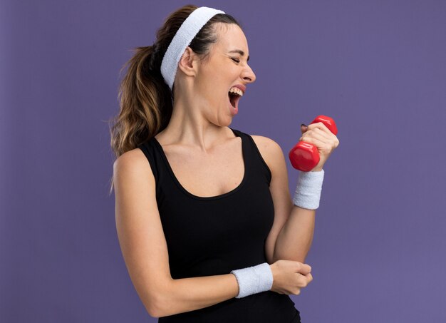 Strained young pretty sporty woman wearing headband and wristbands holding dumbbell putting hand on elbow with closed eyes isolated on purple wall