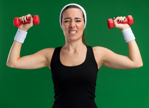Strained young pretty sporty woman wearing headband and wristbands raising dumbbells