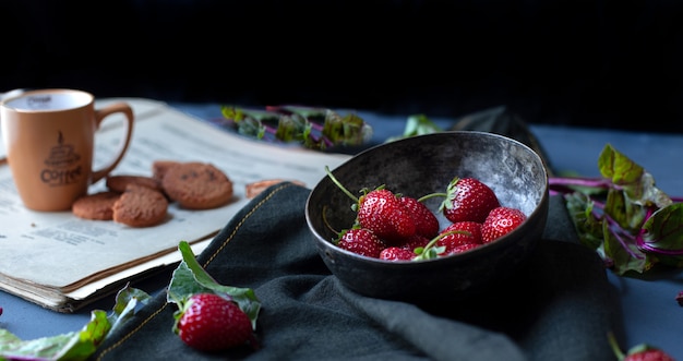 Strawberries inside bowl, cookies and coffee cup on a book paper. 