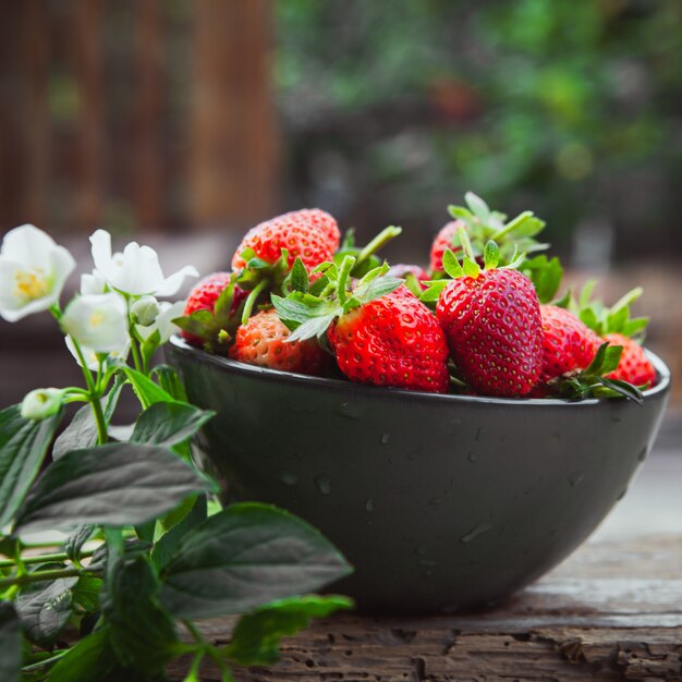 Strawberries with flowers on branch in a bowl on wooden and yard table, side view.