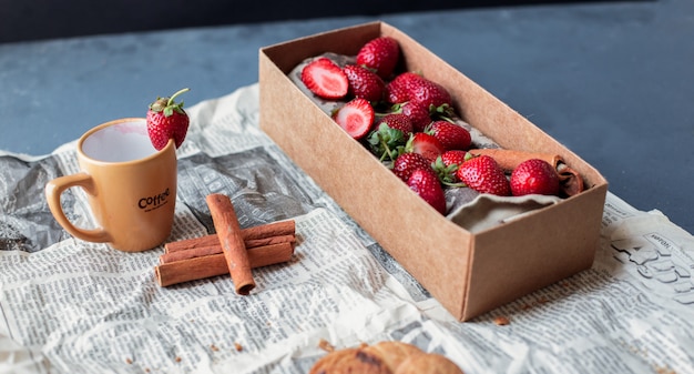 Foto gratuita contenitore di cartone della fragola con la tazza e cinnamons su un giornale.