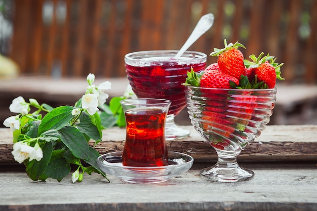 Strawberry jam in a plate with spoon, tea in glass, strawberries, flower branch side view on wooden and yard table