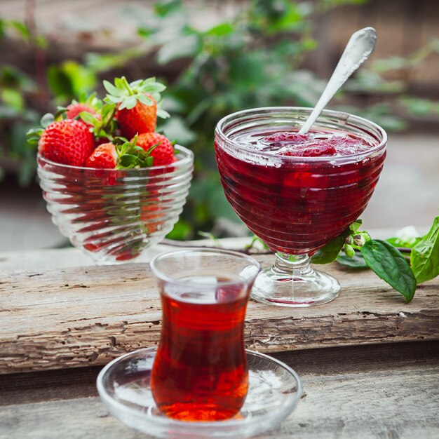 Strawberry jam in a plate with spoon, tea in glass, strawberries, plant close-up on wooden and yard table