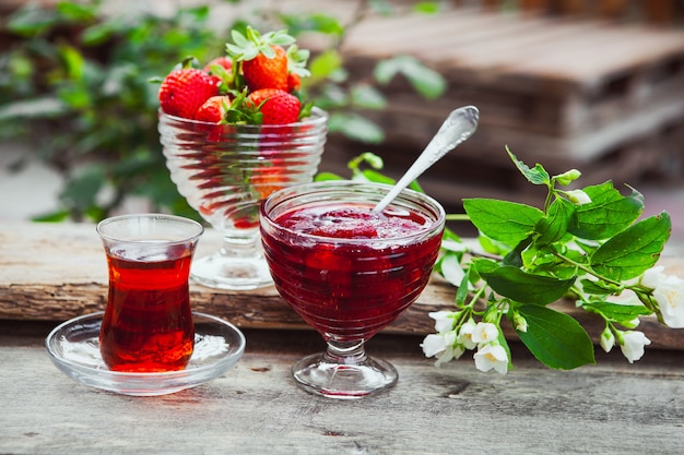 Strawberry jam in a plate with spoon, tea in glass, strawberries, plant side view on wooden and yard table