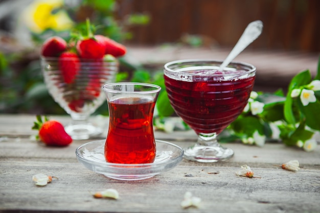 Strawberry jam with flowers on branch, a glass of tea, spoon, strawberries in a plate on wooden and yard table, side view.