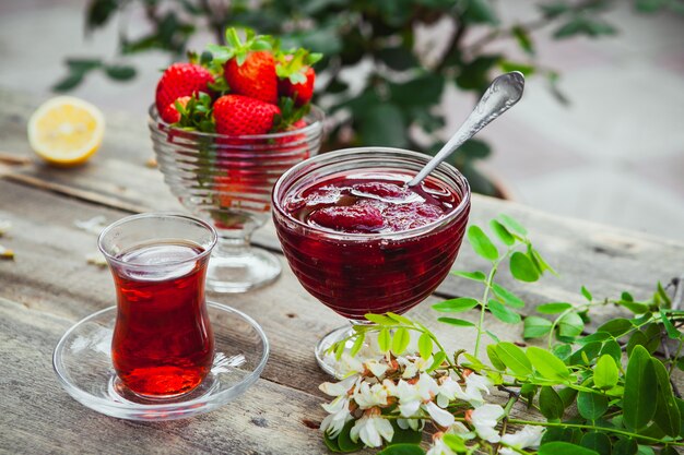 Strawberry jam with spoon, a glass of tea, strawberries, lemon, plants in a plate on wooden and pavement table, high angle view.