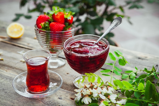 Strawberry jam with spoon, a glass of tea, strawberries, lemon, plants in a plate on wooden and pavement table, high angle view.