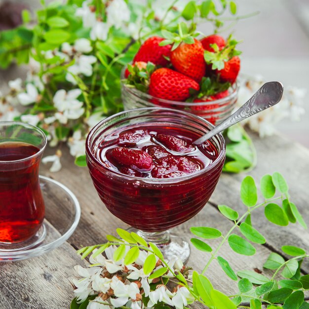 Strawberry jam with spoon, a glass of tea, strawberries, plant in a plate on wooden and pavement table, close-up.