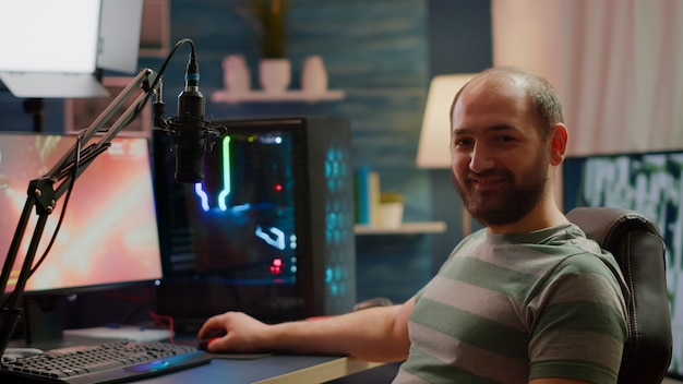 Streamer man looking at camera smiling while streaming videogames using stream chat. Pro cyber videogamer playing space shooter video game at RGB powerful personal computer in gaming home studio