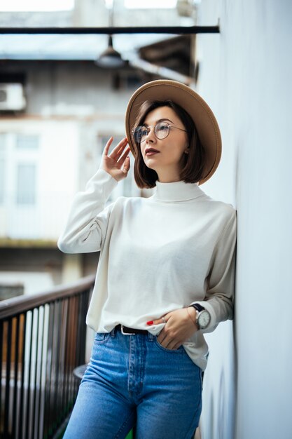 Street fashion interested woman wearing hat, blue jeans, wide hat and transperent glasses on balcony