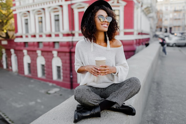 Foto gratuita look alla moda di strada. ragazza nera alla moda che si siede sul ponte e che tiene tazza di caffè