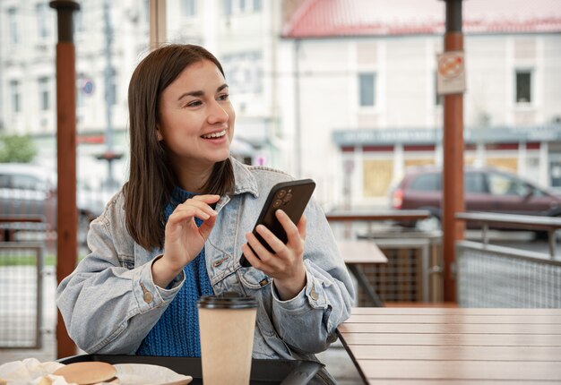 Street portrait of a cheerful young woman on a cafe terrace, holding a phone.