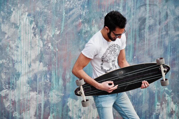 Street style arab man in eyeglasses with longboard posed against colored wall like he play guitar