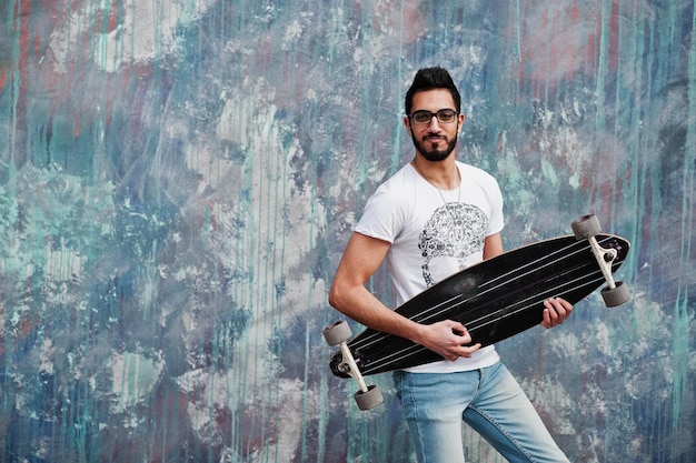 Street style arab man in eyeglasses with longboard posed against colored wall like he play guitar