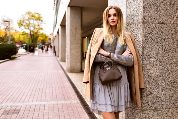 Street style fashion portrait of elegant glamour blonde woman posing on the European street near shopping center, elegant dress sweater and cashmere coat, toned colors, spring time.