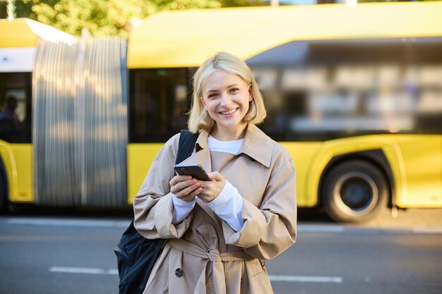 Foto gratuita street style shot di una giovane donna bionda sorridente che tiene in mano un telefono cellulare in piedi per strada e guardando