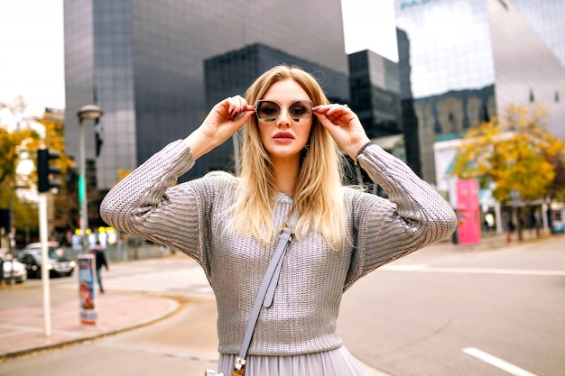 Street stylish portrait of blonde woman wearing glamour grey outfit at put hand to her sunglasses, business center area.