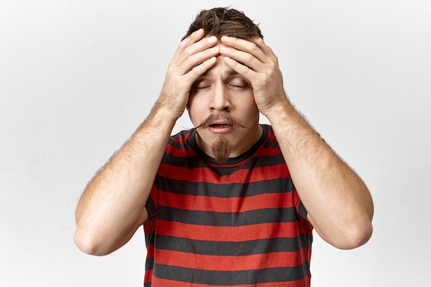 Stress, problems and negative emotions concept. Picture of fashionable young unshaven man in striped t-shirt closing eyes and massaging forehead, suffering from headache, trying to concentrate
