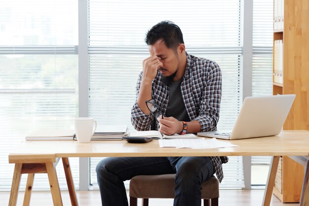 Stressed Asian man sitting at table with laptop and documents and rubbing forehead