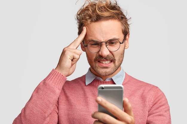 Stressed bearded man keeps fore finger on temple, has displeased look into smart phone, frowns face in displeasure