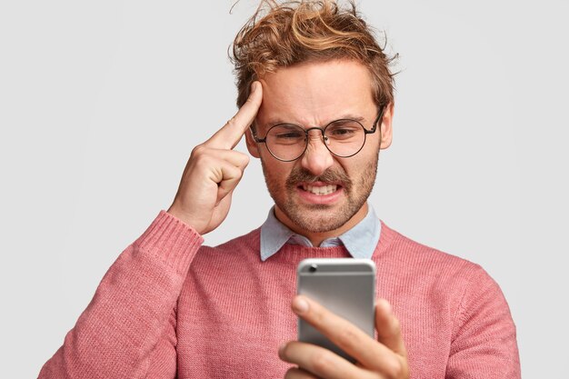 Подчеркнутый бородатый мужчина держит указательный палец на виске, недовольно смотрит в смартфон, недовольно хмурится
