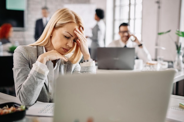 스트레스를 받은 여성 사업가는 고통에 머리를 잡고 직장에서 두통을 겪고 있습니다. 배경에 사람들이 있습니다