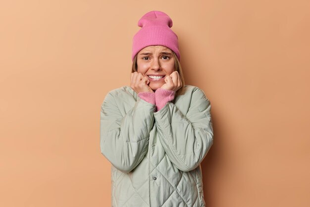 Stressed concerned woman clenches teeth looks anxious and bothered afraids of something terrifying trembles from fear wears hat and jacket isolated over beige background Negative emotions reactions