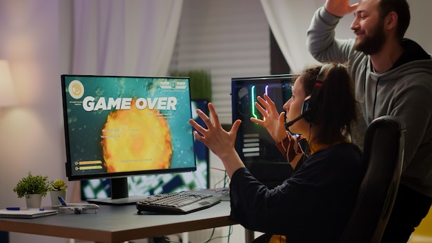 Подчеркнутая пара геймеров проигрывает космический шутер, играя на мощном компьютере с RGB-подсветкой во время потокового онлайн-соревнования. Профессиональная кибер-женщина с гарнитурой выступает из дома во время виртуального турнира