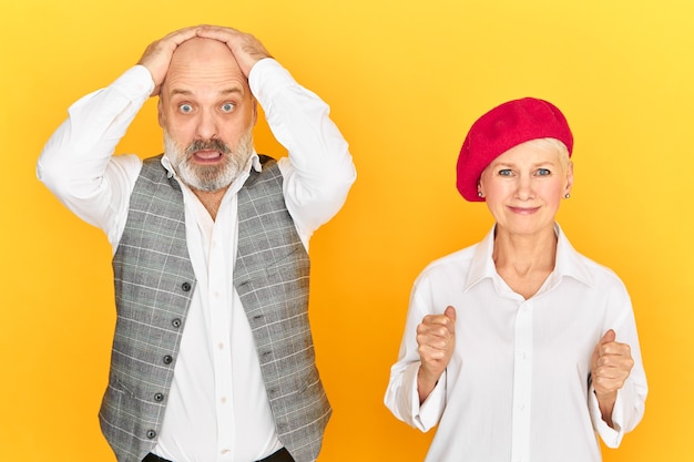 Stressed elderly male with beard holding hands on head and staring at camera with full disbelief, falling into panic, his wife trying to support him