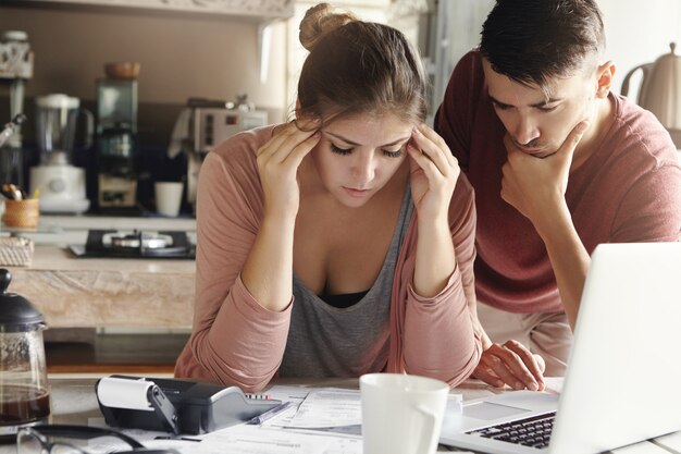 ストレスのたまった女性は、金融危機の緊張に耐えられず、寺院を圧迫し、紙幣の山、ノートパソコン、電卓を備えた台所のテーブルに座っています。解決策を見つけようとする彼女のそばにいる夫