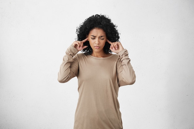 Free photo stressed frustrated young dark-skinned woman wearing beige long-sleeved top plugging her ears