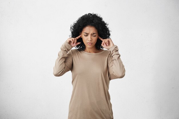 Stressed frustrated young dark-skinned woman wearing beige long-sleeved top plugging her ears