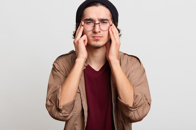 Stressed handsome youngster touching his temples with both hands, feeling headache, trying to reduce it, looks upset. Black haired slim model wears black hat, red and beige shirts, spectacles.