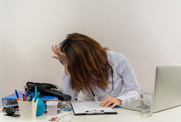 Stressed middle-aged female doctor wearing medical robe and stethoscope sitting at desk with medical tools clipboard and laptop putting hands on head and on desk isolated