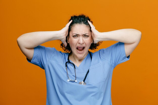 Stressed middleaged female doctor wearing uniform and stethoscope around her neck keeping hands on head looking at camera screaming isolated on orange background