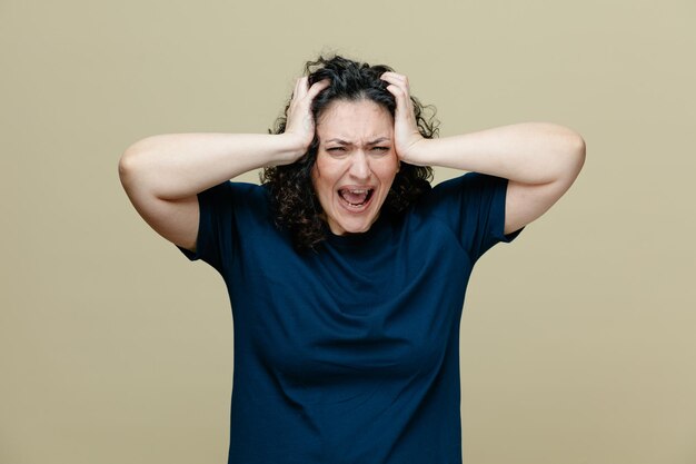 Stressed middleaged woman wearing tshirt keeping hands on head looking at camera screaming isolated on olive green background