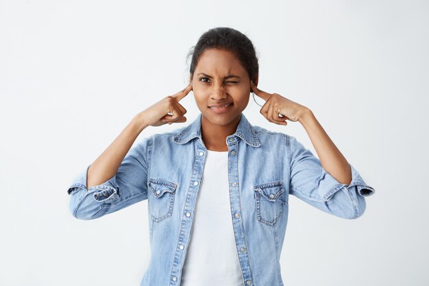 Stressed nervous young dark-skinned woman with black hair closing eyes and plugging ears with fingers, annoyed with loud sound or music coming from neighbour's flat. Stop making this noise