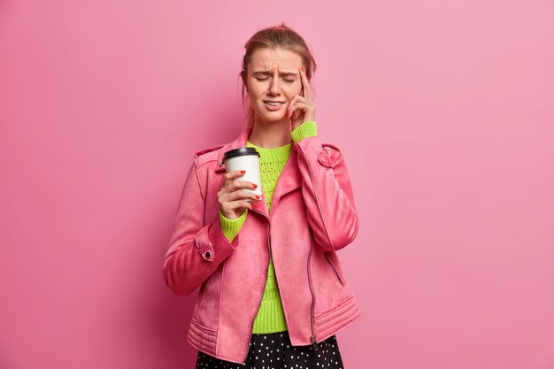 Stressed overworked woman touches temples, has unbearable headache, drinks takeaway coffee, wears pink jacket, closes eyes to relieve pain, poses indoor. People, lifestyle