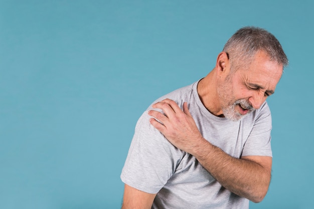 Stressed senior man with shoulder pain on blue backdrop