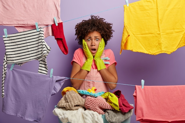 Stressed shocked woman with Afro hairstyle does laundry at home, hangs wet clean linen on clothesline, wears casual clothes and rubber gloves, stunned to have much work. Emotional housekeeper