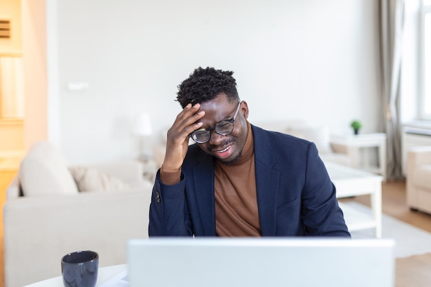 Stressato uomo afroamericano stanco che tocca le tempie che soffrono di mal di testa dopo lunghe ore di lavoro oberati di lavoro uomo d'affari sopraffatto seduto alla scrivania che si sente male