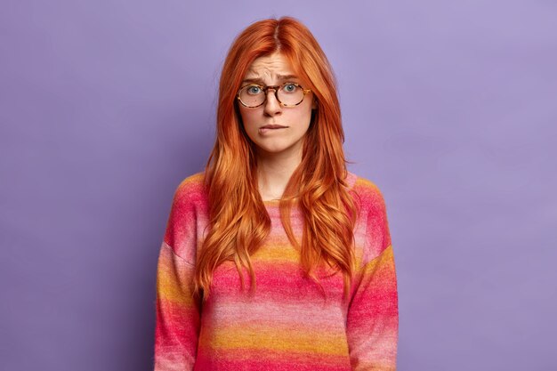 Stressed worried ginger woman bites lips nervously being anxious about troubles or problem wears transparent glasses and sweater.