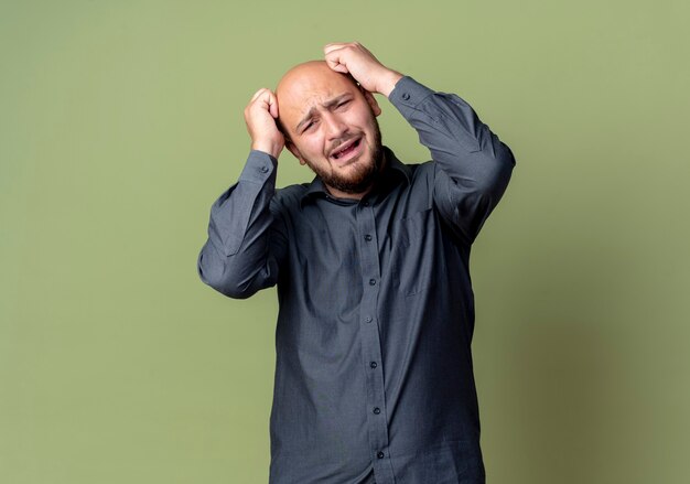 Stressed young bald call center man putting hands on head isolated on olive green wall