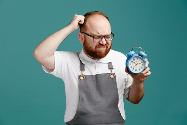 Стрессовый молодой парикмахер в униформе и очках, показывающий будильник, касающийся головы кулаком с закрытыми глазами на синем фоне