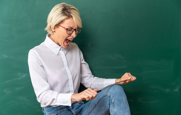 Stressed young blonde female teacher wearing glasses in classroom standing in profile view in front of chalkboard screaming breaking pointer stick on leg with closed eyes