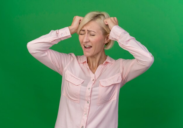 Stressed young blonde slavic woman touching head with fists with closed eyes isolated on green background